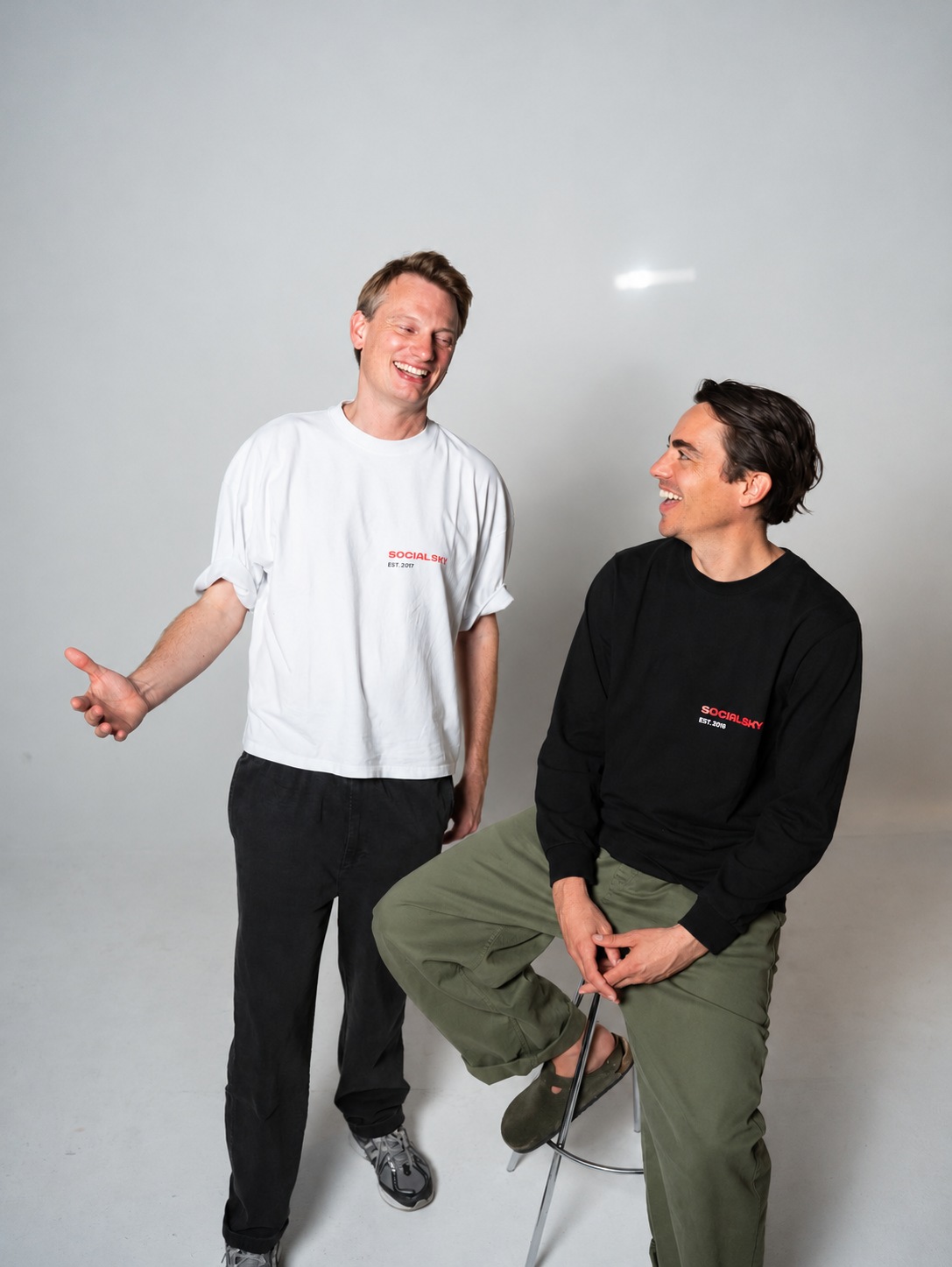 Deux hommes souriants, l’un debout, l’autre assis sur un tabouret, devant un fond de studio gris